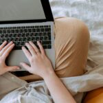 Anonymous woman in casual clothes sitting on sofa and browsing laptop with blank screen while working on project on bed