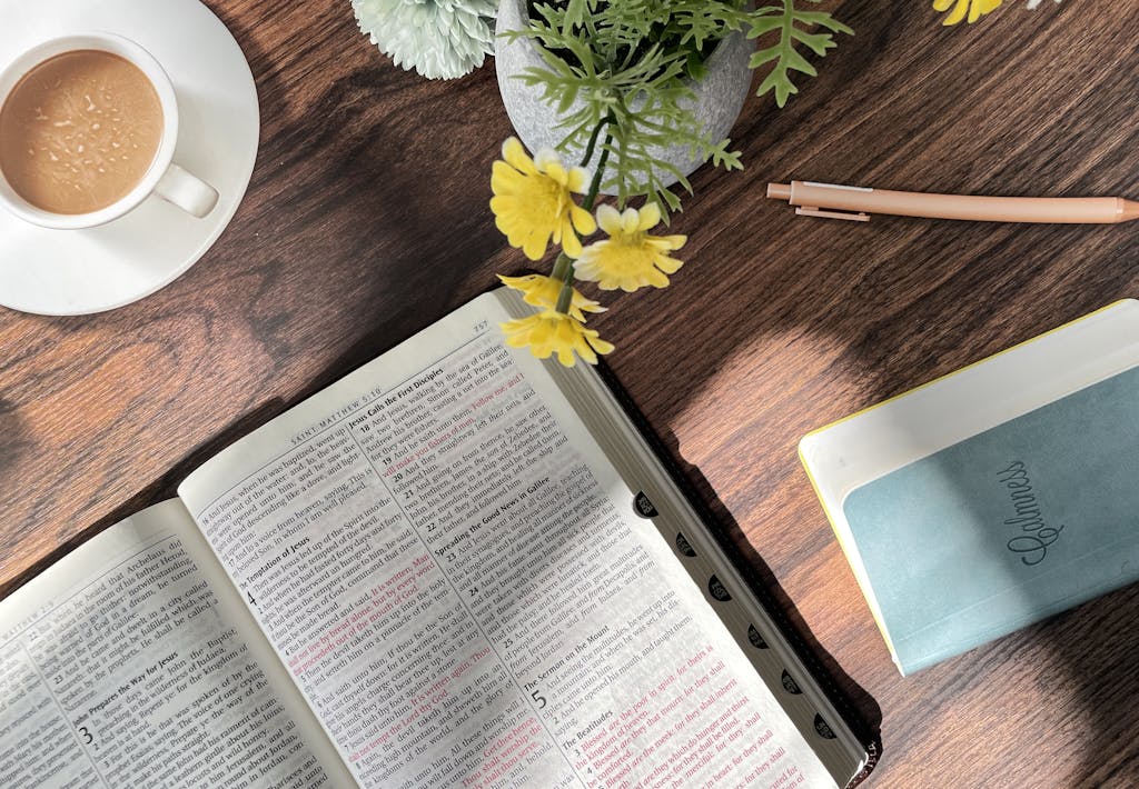 Flat lay showing an open Bible, coffee cup, notebook, and flowers on a wooden table in sunlight.