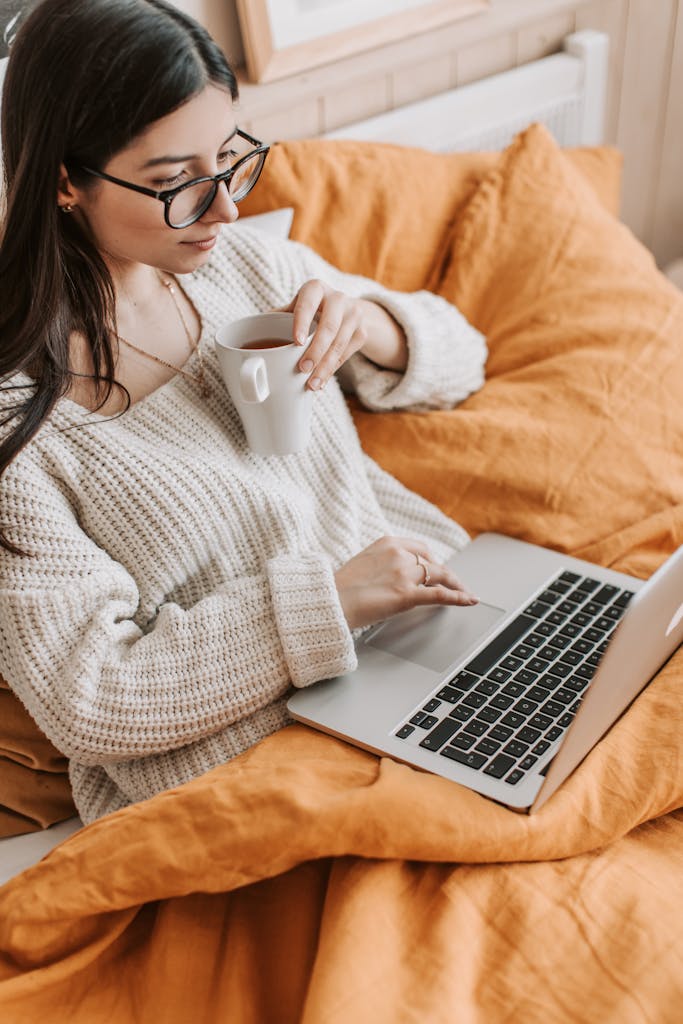 Woman enjoying coffee while working on a laptop in a cozy bedroom setting.