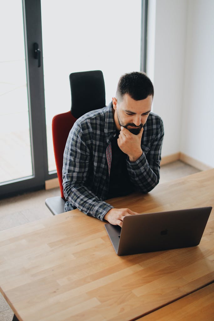 Young man with beard working on laptop at desk in a modern office setting.