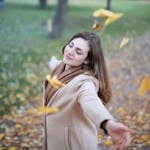 Young woman with arms outstretched, delighting in fallen leaves in a Paris park during autumn daytime.