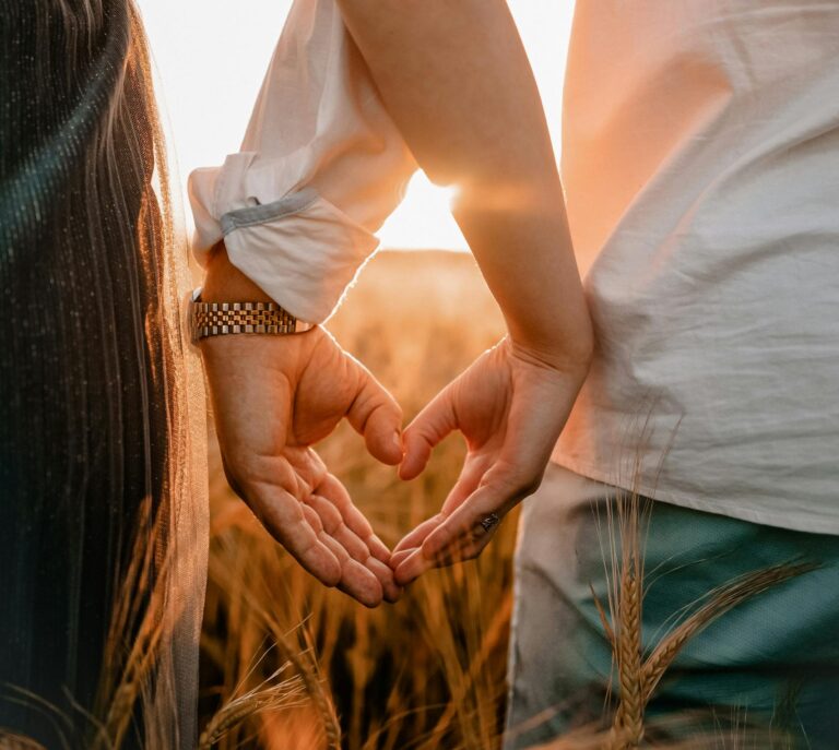 Couple standing in field create heart shape with hands at sunset, symbolizing love.