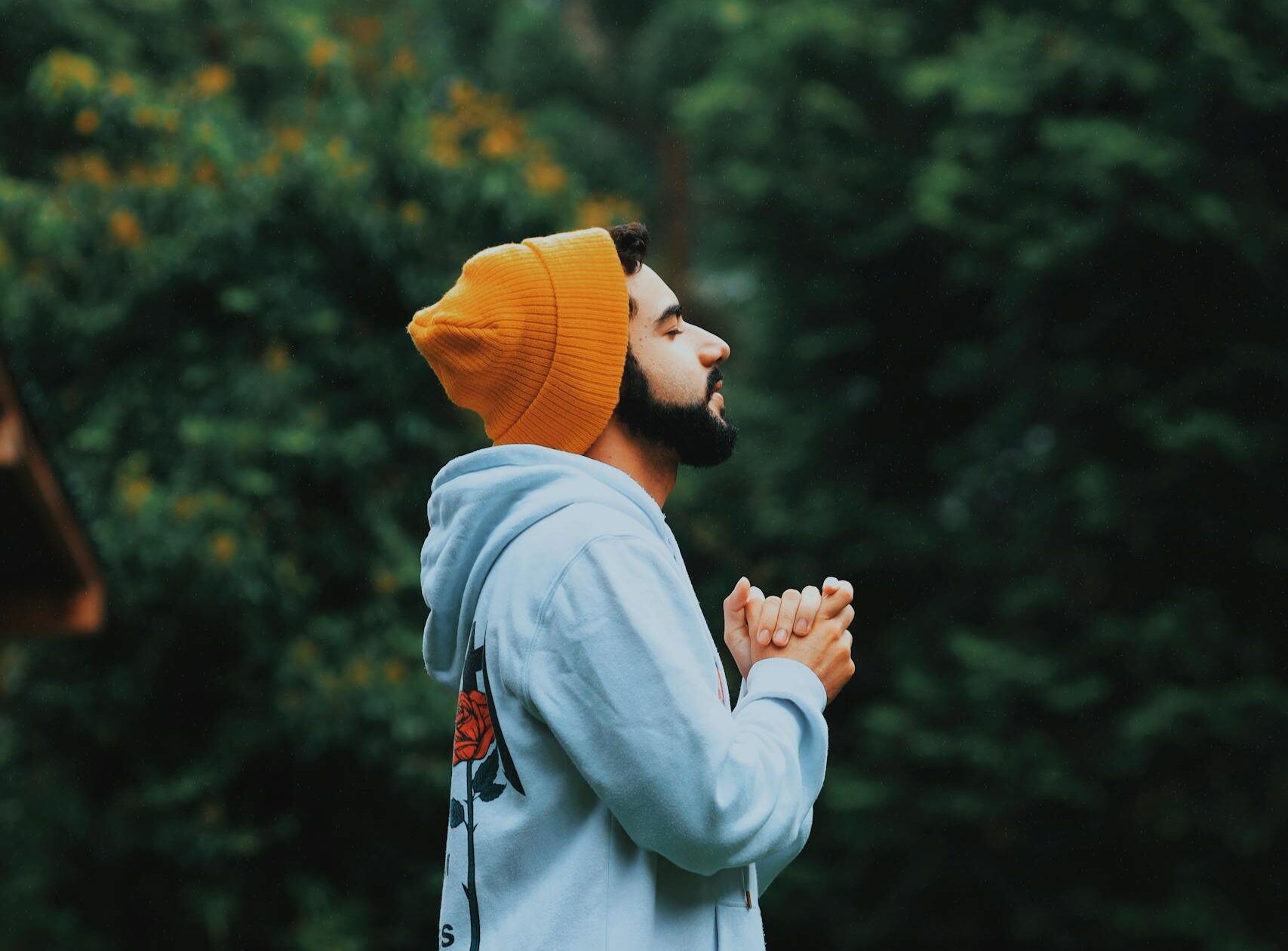 A man in a beanie and hoodie deeply praying outside in a lush green forest.