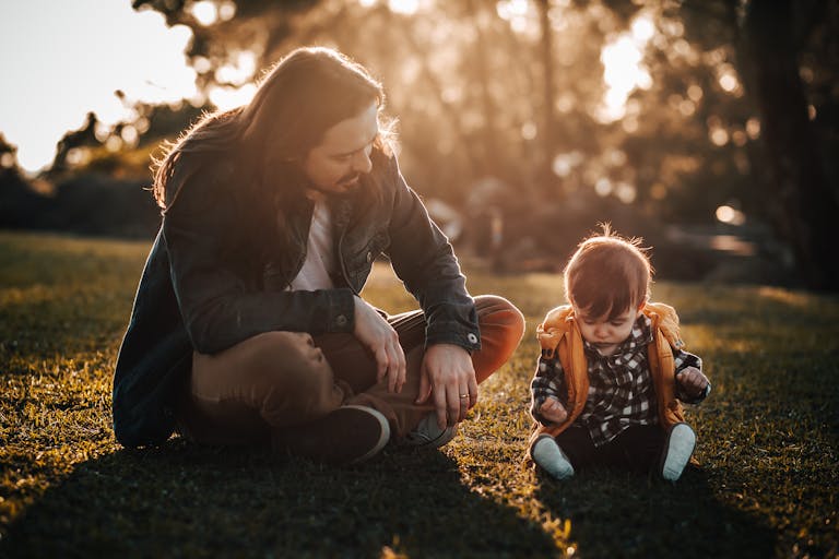 Full body of father and son sitting together on grassy ground in park while looking down in summer evening
