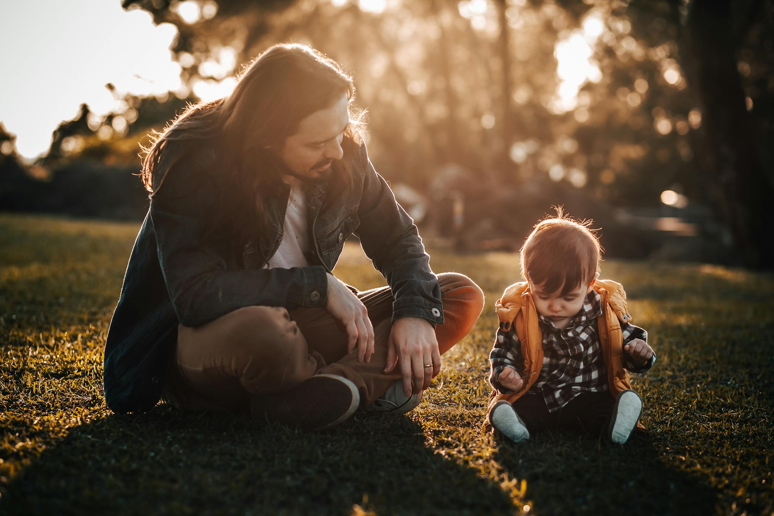 Full body of father and son sitting together on grassy ground in park while looking down in summer evening
