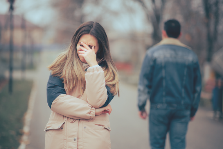 A heartbroken woman covers her face in tears as her husband walks away, a picture of the pain of betrayal trauma in marriage.