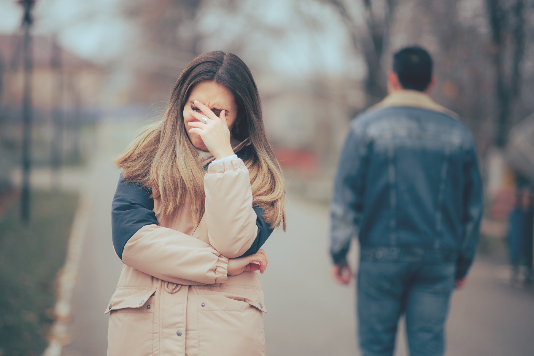 A heartbroken woman covers her face in tears as her husband walks away, a picture of the pain of betrayal trauma in marriage.